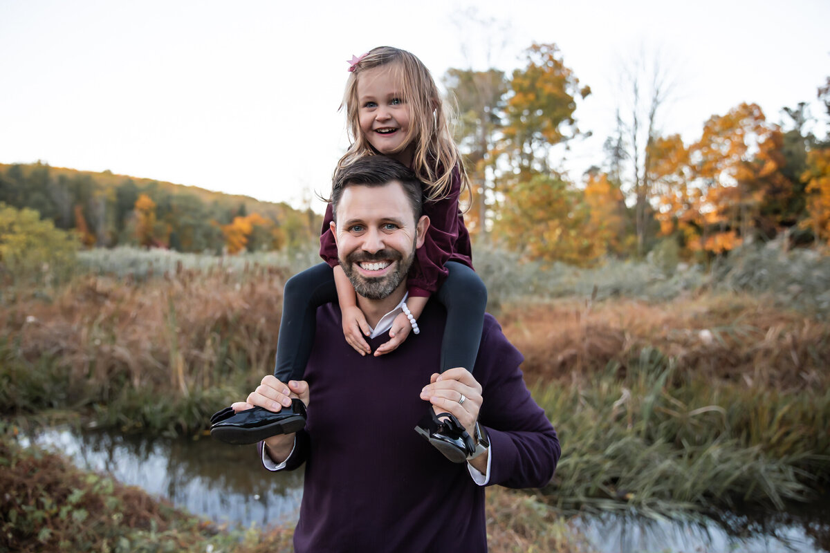 A Bel Air, Maryland father holds his daughter on his shoulders smiling during their fall lifestyle family picture at Harford Glen Park.