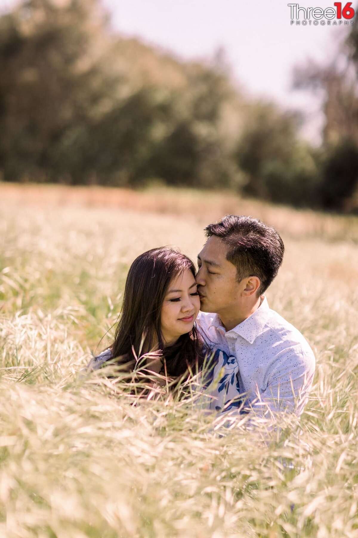 Romantic gesture as Groom to be kisses his fiance's cheek while sitting in tall grass