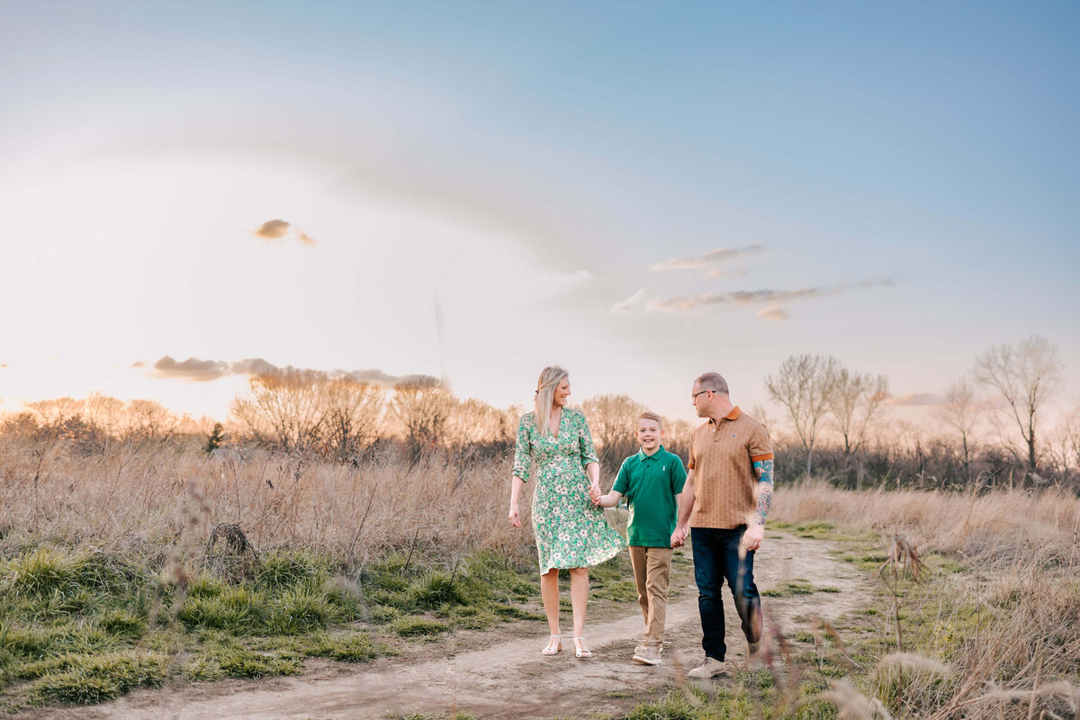 family of 3 walking hand in hand with thier son between them, walking on a curvy dirt path at sunset