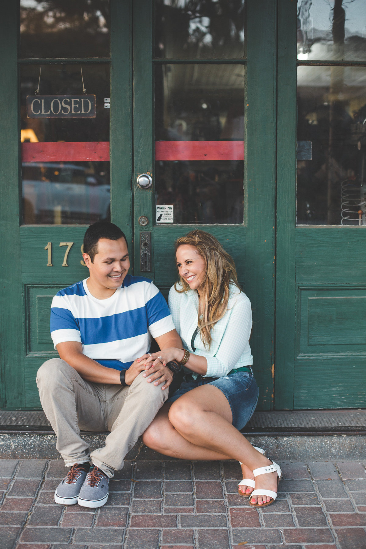 Engaged couple sitting in on cobblestone path in front from green door holding hands.