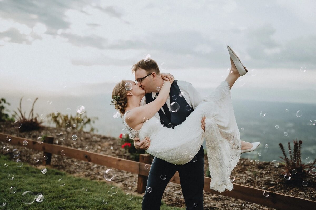 A bride and groom share a kiss amidst floating bubbles, with a scenic ocean view in the background