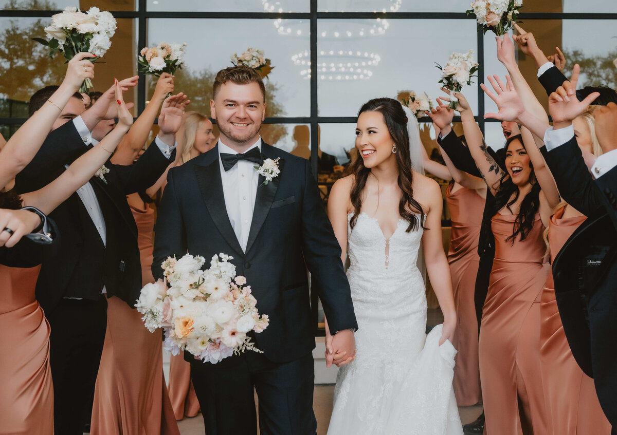 Bride and groom walk hand in hand smiling as wedding guests in satin dresses and tuxedos cheer and raise bouquets in celebration.