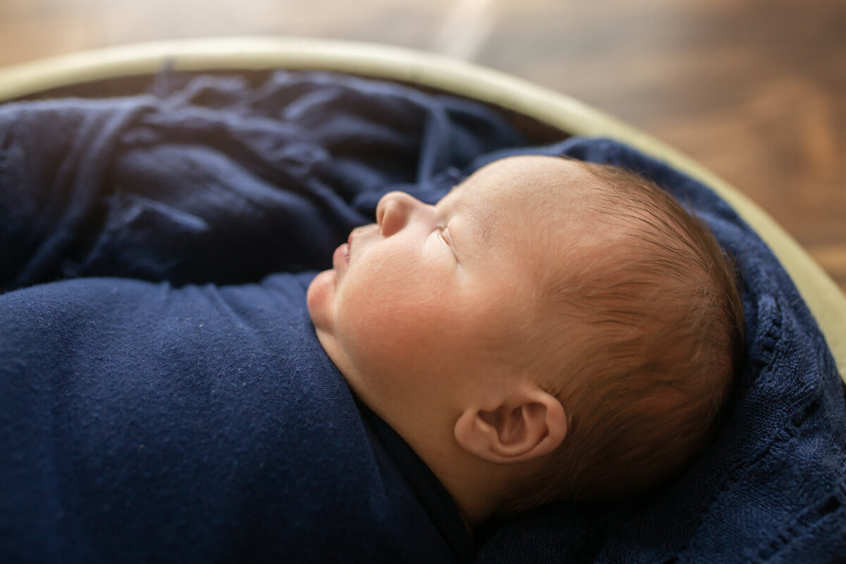 A newborn baby boy sleeps in a basket wrapped in a navy blanket