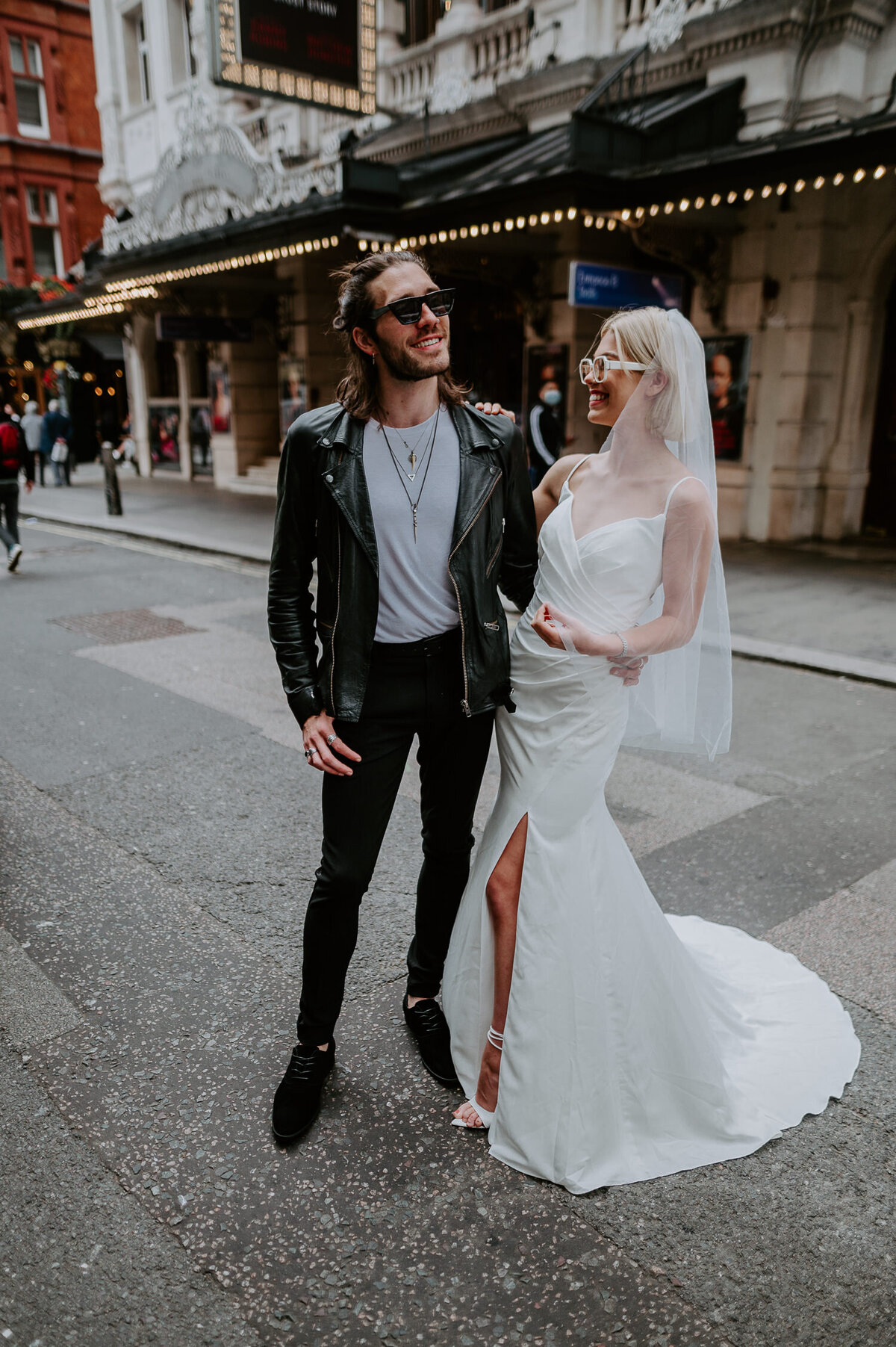 A bride and groom stand in the west end of London outside a theatre. The groom is wearing glasses, has long hair and is also wearing a leather jacket. The bride is also wearing sun glasses and has a wedding dress on designed by Justin Alexander.