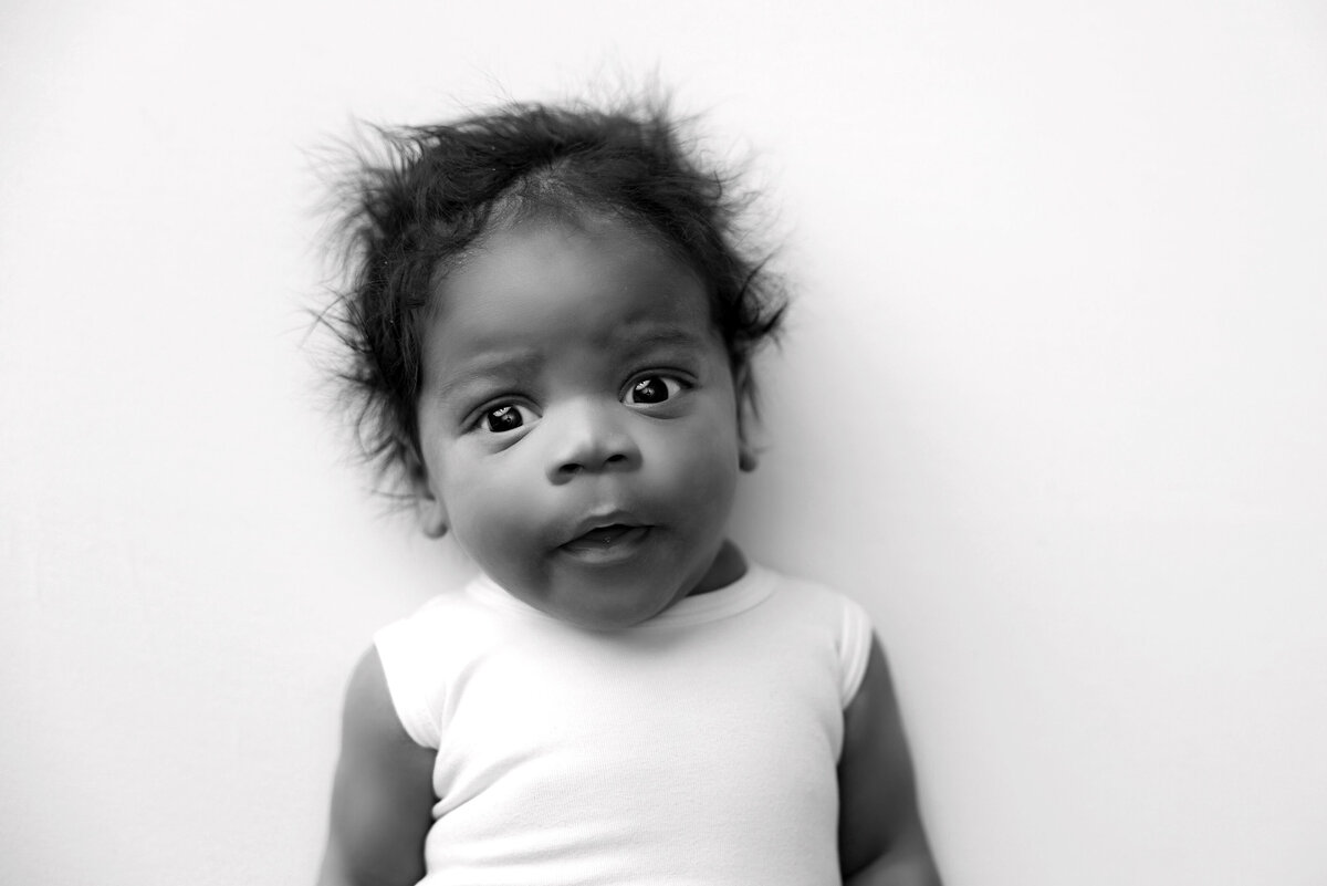 Black and white image of a baby with curly hair, wearing a white sleeveless outfit. Captured by a Jacksonville newborn photographer, the baby has a curious expression and is lying on a light-colored surface.
