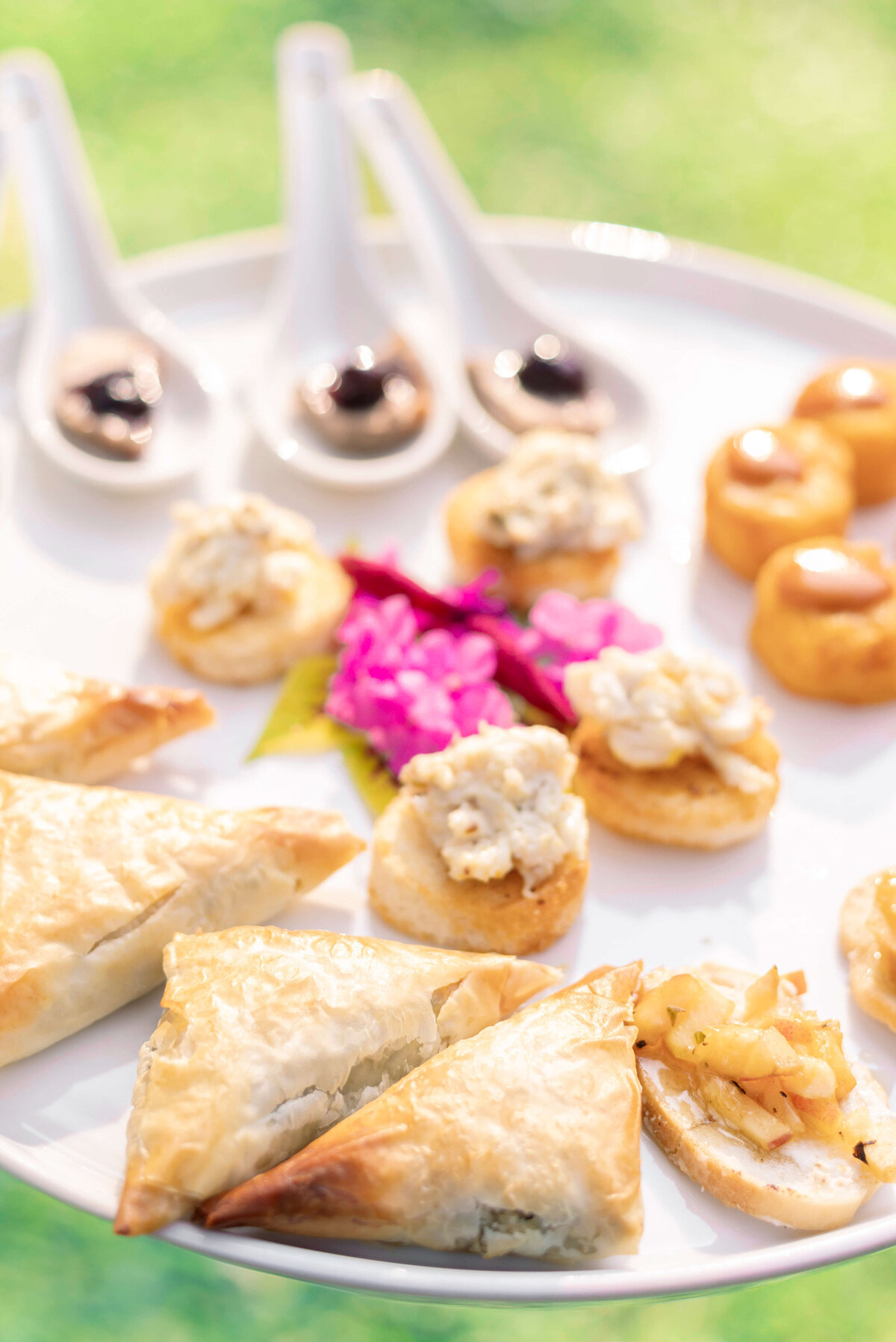 A beautifully arranged platter featuring various appetizers: flaky pastries, crostini with creamy toppings, and small round bread bites. Behind them, four porcelain spoons hold dark sauces. A pink flower garnish adds a pop of color.