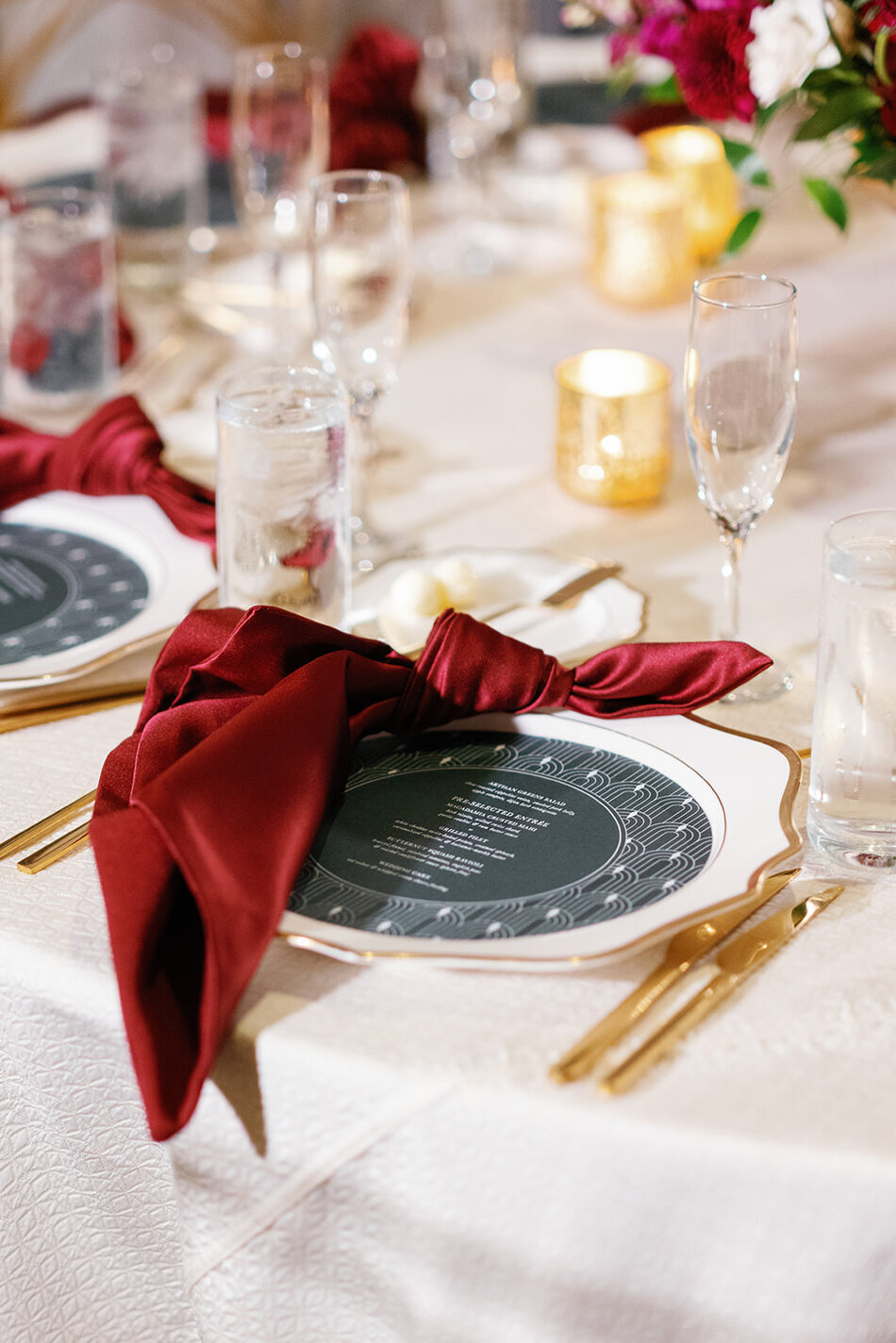 Stylish table arrangement at the Don Cesar Hotel in St. Pete Beach, FL, showcasing white linens, gold edge chargers with scalloped details, scarlet red knotted napkins, and round green wedding menus perfectly positioned inside the chargers.