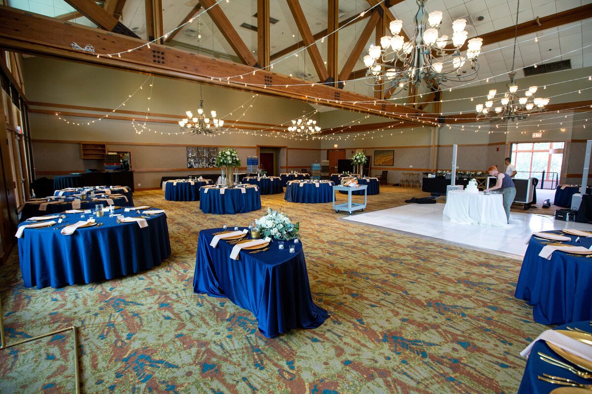 Elegant event hall with blue tablecloths, set tables, string lights, and a large chandelier, with attendees in the background, designed by a top wedding planner in Des Moines.