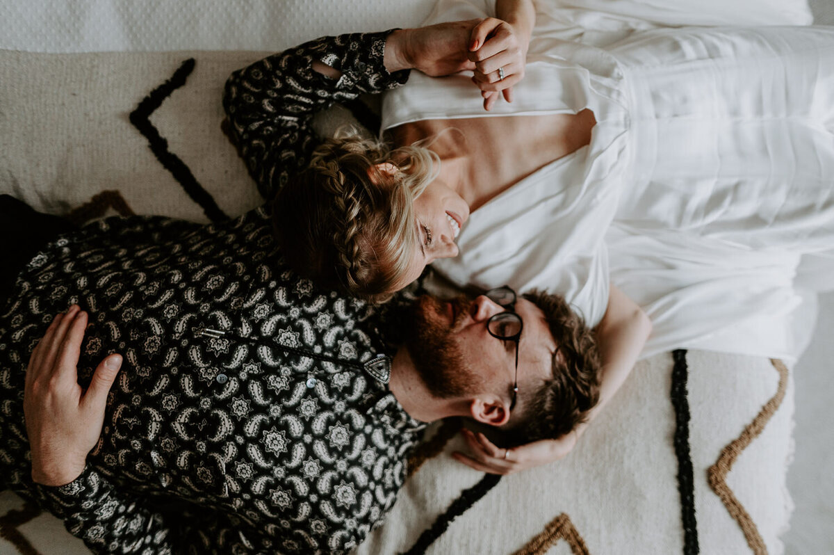 A bride and groom lay on their bed dressed up in their wedding outfits. The bride is wearing a v cut bohemian wedding dress and the groom is wearing a black and white patterned shirt.