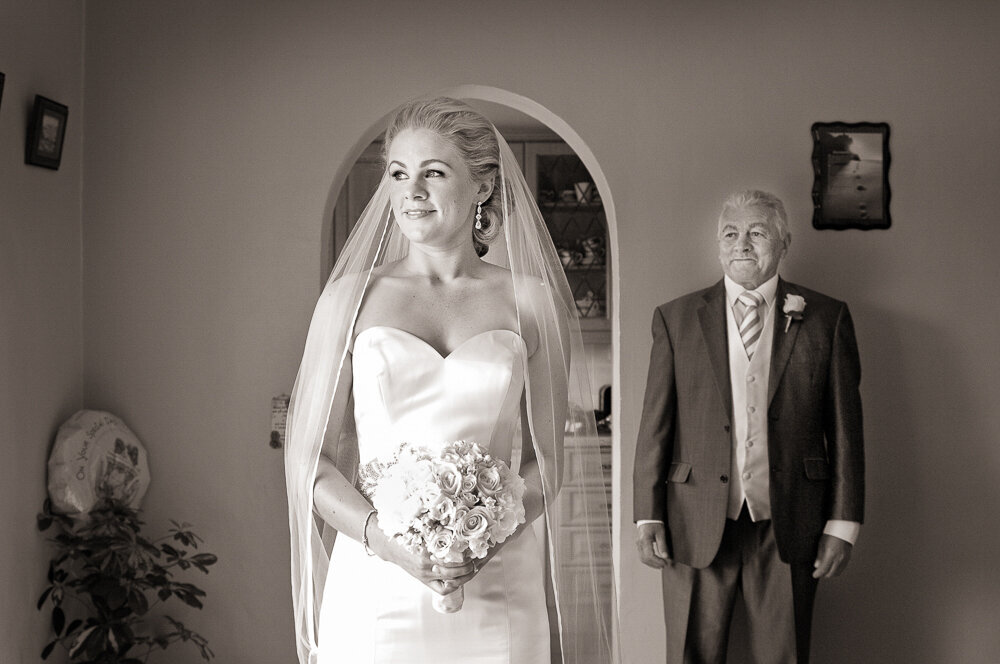 blonde bride wearing a mermaid style wedding dress and long veil looking out window while the father of the bride looks at her