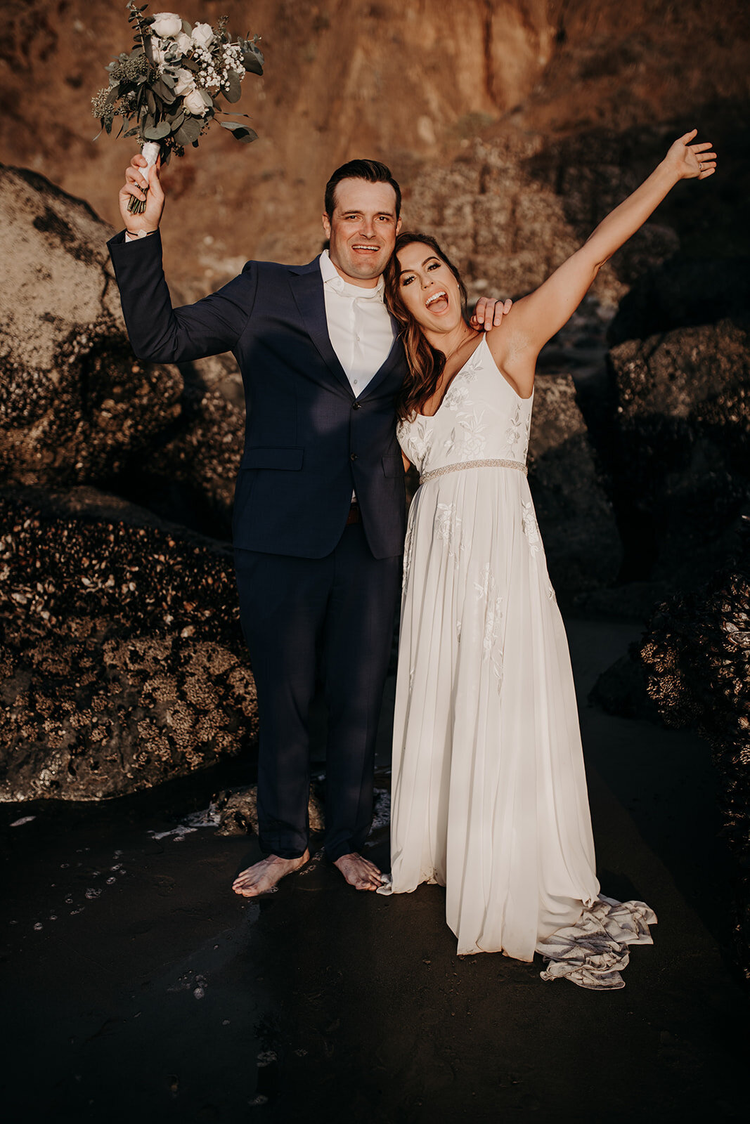 Bride & Groom cheering as they stand barefoot on a rocky beach