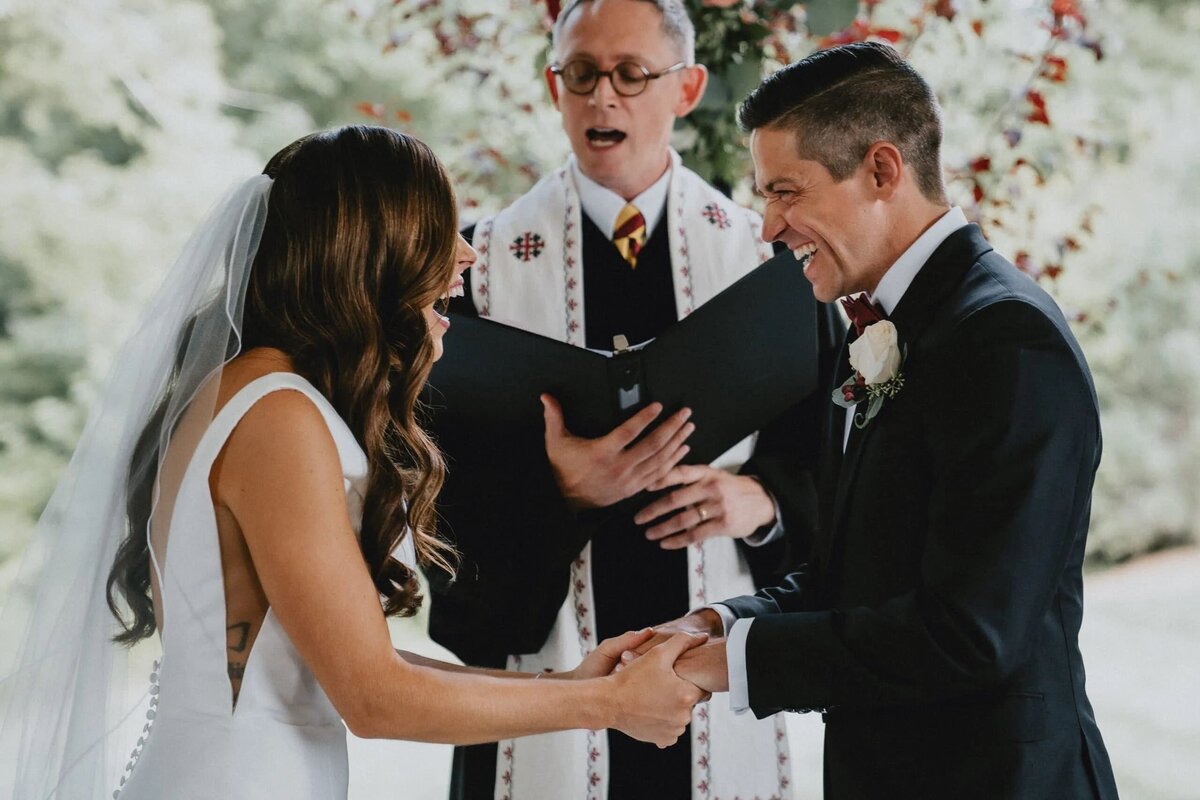 The bride and groom hold hands at the altar, with a smiling officiant and bridesmaids in the background of this heartwarming ceremony.