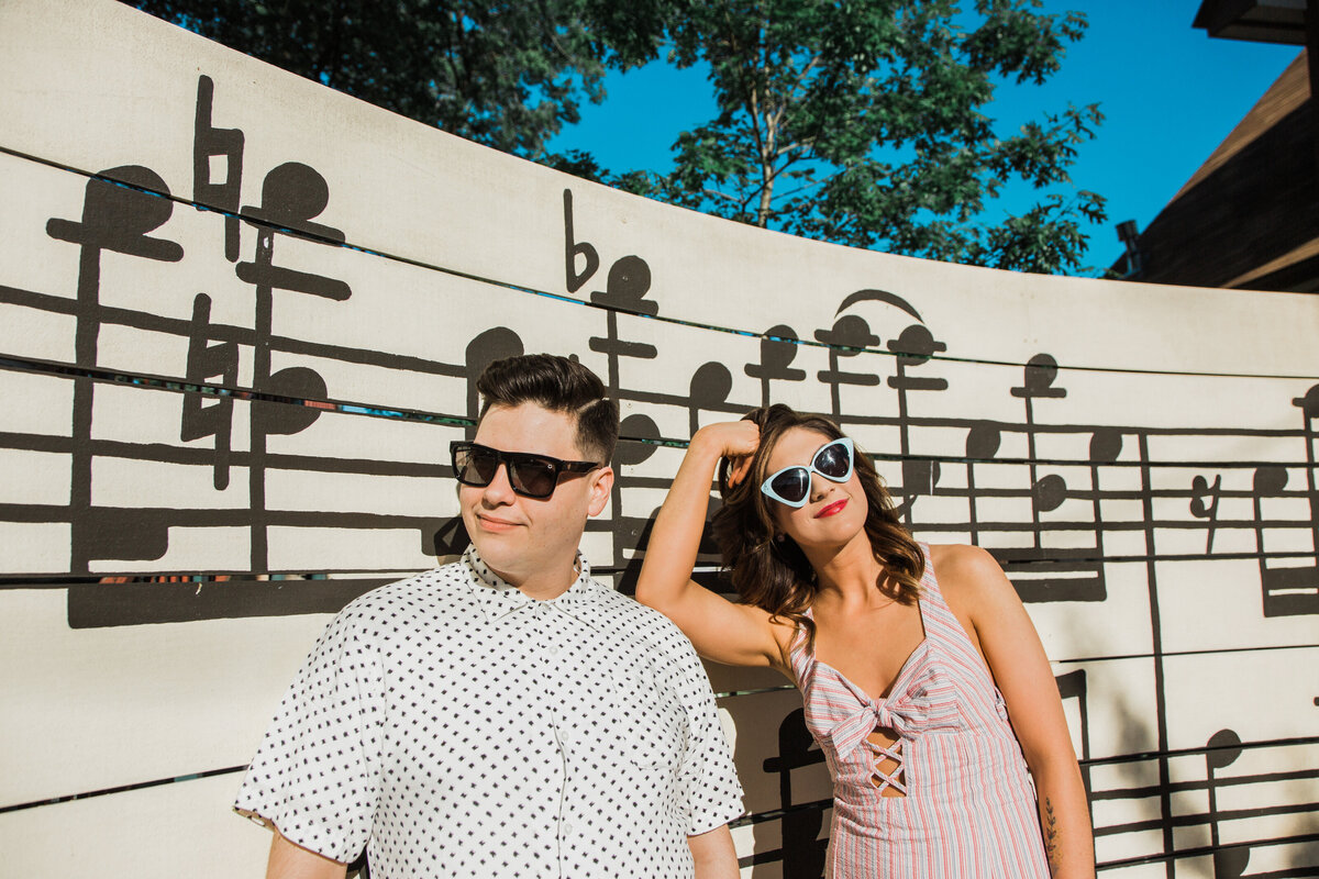A couple casually posing in front of a musical mural during their engagement photoshoot in the Bishop Arts District in Dallas, Texas. The woman on the right is wearing a colorful, stripped sundress and retro blue sunglasses. The man on the left is wearing a short sleeve dress shirt and black sunglasses. The mural behind them depicts a portion of the musical score of Scott Joplin's Maple Leaf Rag.