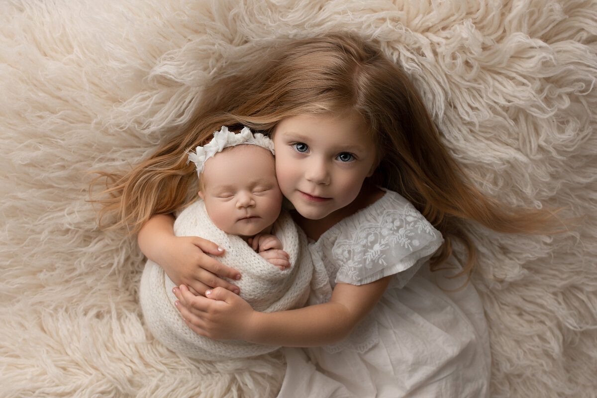 Fine art newborn photos captured by New Jersey's best newborn photographer Katie Marshall. Aerial image. Big sister in a cream dress has her arms wrapped around her baby sister who is swaddled in white with a matching headband. The baby is sleeping and her hands are peeking out of the swaddle. Big sister has her arms wrapped around baby and resting her cheek against the baby's head.