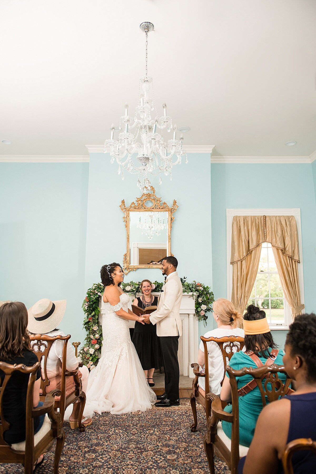 The bride and groom get married in the front parlor at Ravenswood. The walls are light blue with long gold curtains around the window and an elaborate gold mirror above the mantle. There is a crystal chandelier hanging about the bride and groom. The white mantle is decorated with a lush garland of greenery and flowers. Guests sit in white chairs as they watch the ceremony. The bride is wearing a fit and flare off the shoulder wedding dress with a long train. The groom is wearing a white tuxedo jacket with black pants. The officiant, holding a leather bound book, is wearing a black dress.