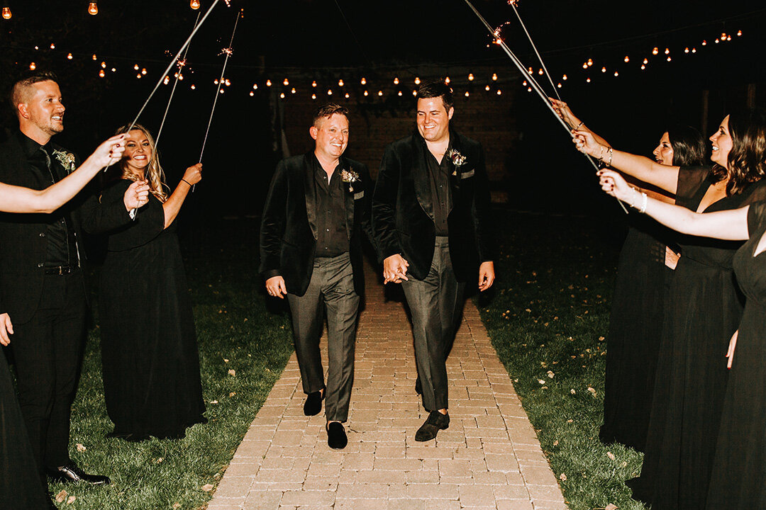 Two grooms wearing black tuxedos walk down a walkway surrounded by friends holding sparklers above as they walk.