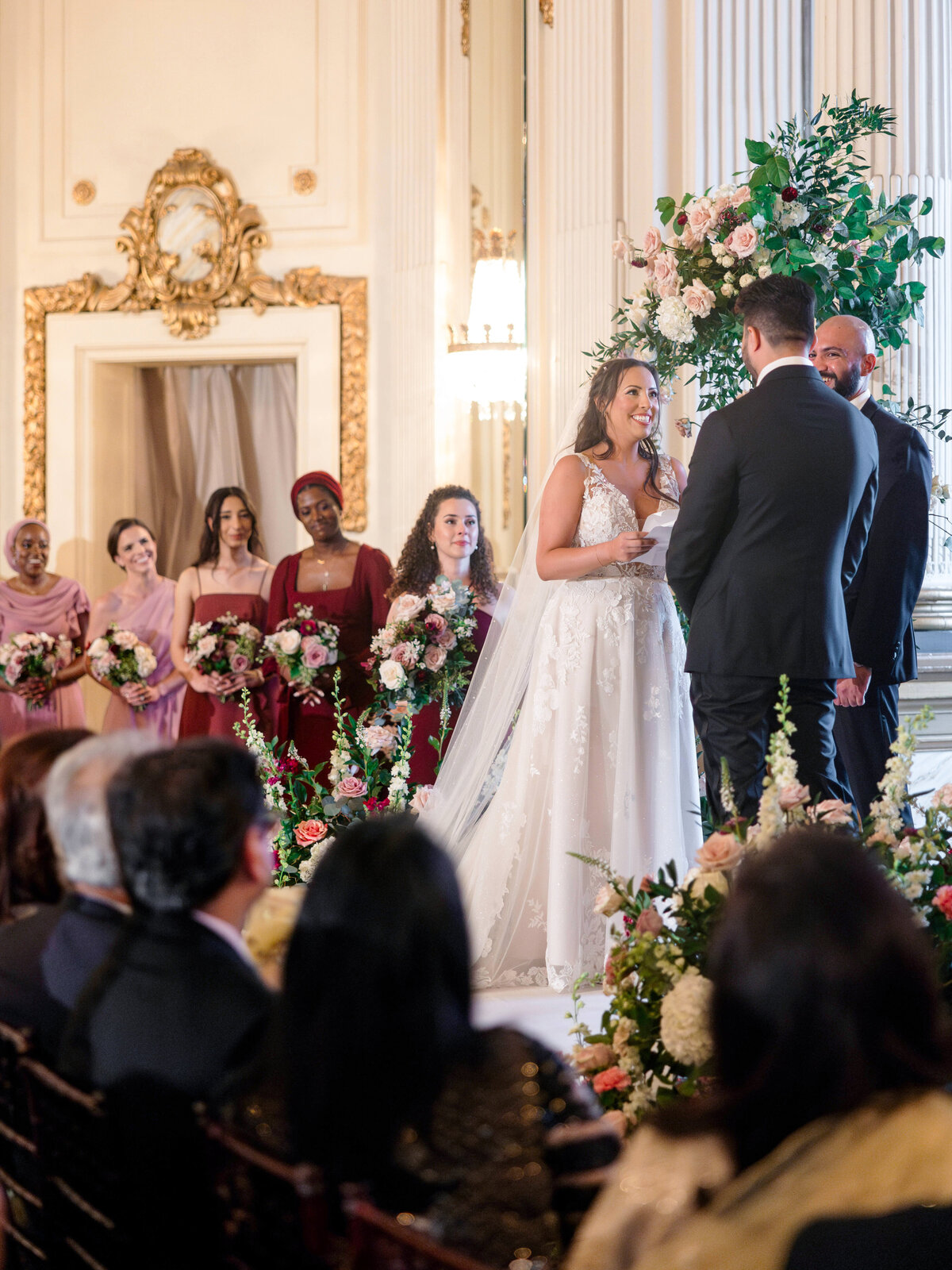 A bride and groom stand at an indoor wedding ceremony. The bride, wearing a white gown, smiles at the groom, who wears a black suit. Bridesmaids in pink dresses hold bouquets and stand beside them. Floral arrangements decorate the elegant room.