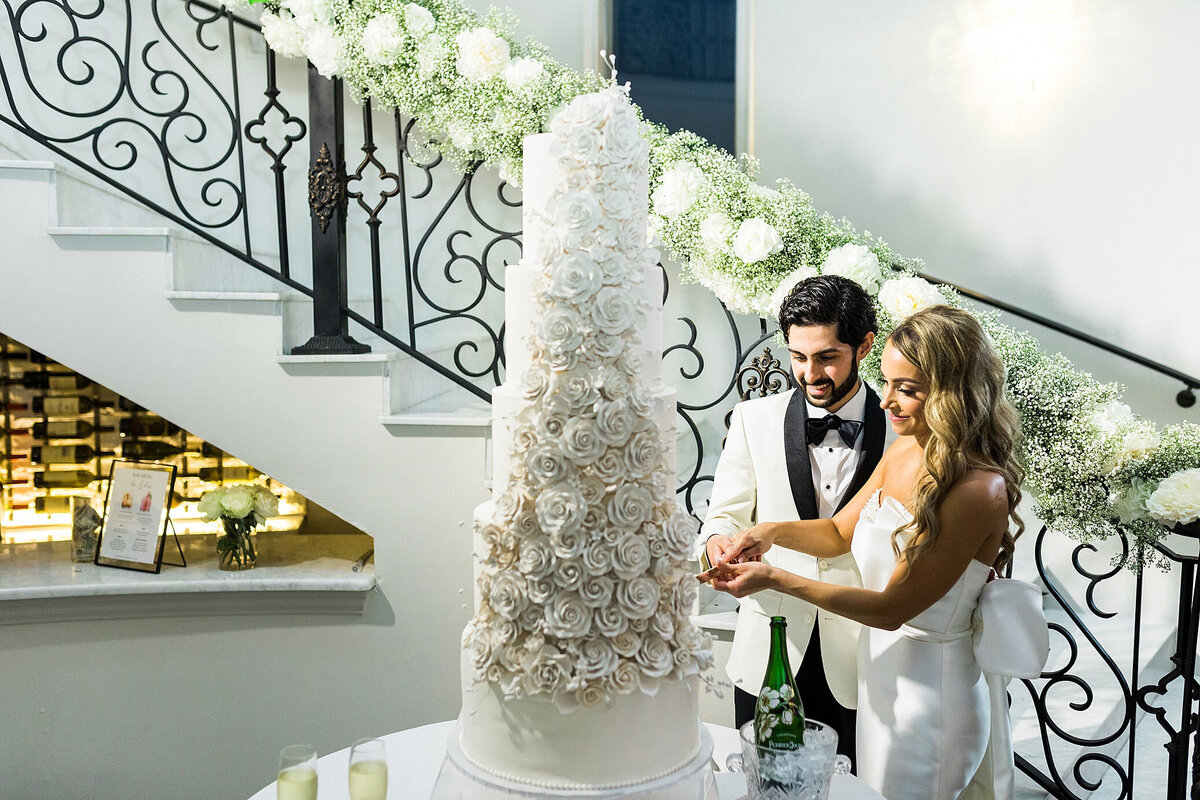 Couple cutting 4 layer tall all white wedding cake with sugar flowers and floral on the staircase
