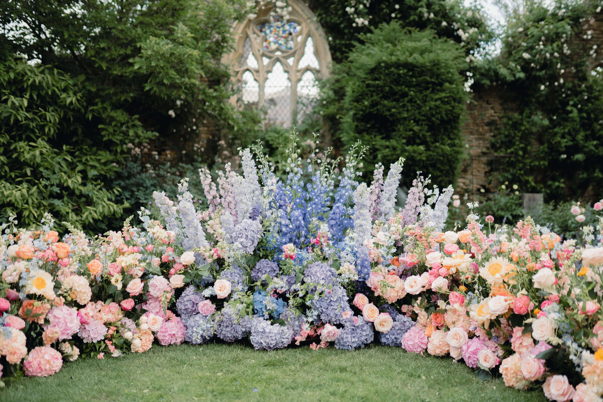 pink and purple flowers in a curved floral wall backdrop to a wedding ceremony in the gardens of a romantic cotswold wedding venue with a church ruin behind