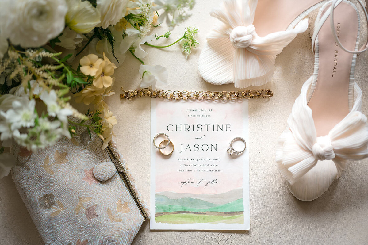 A wedding invitation, two rings, a pair of white shoes, and a floral arrangement laid out on a textured surface.