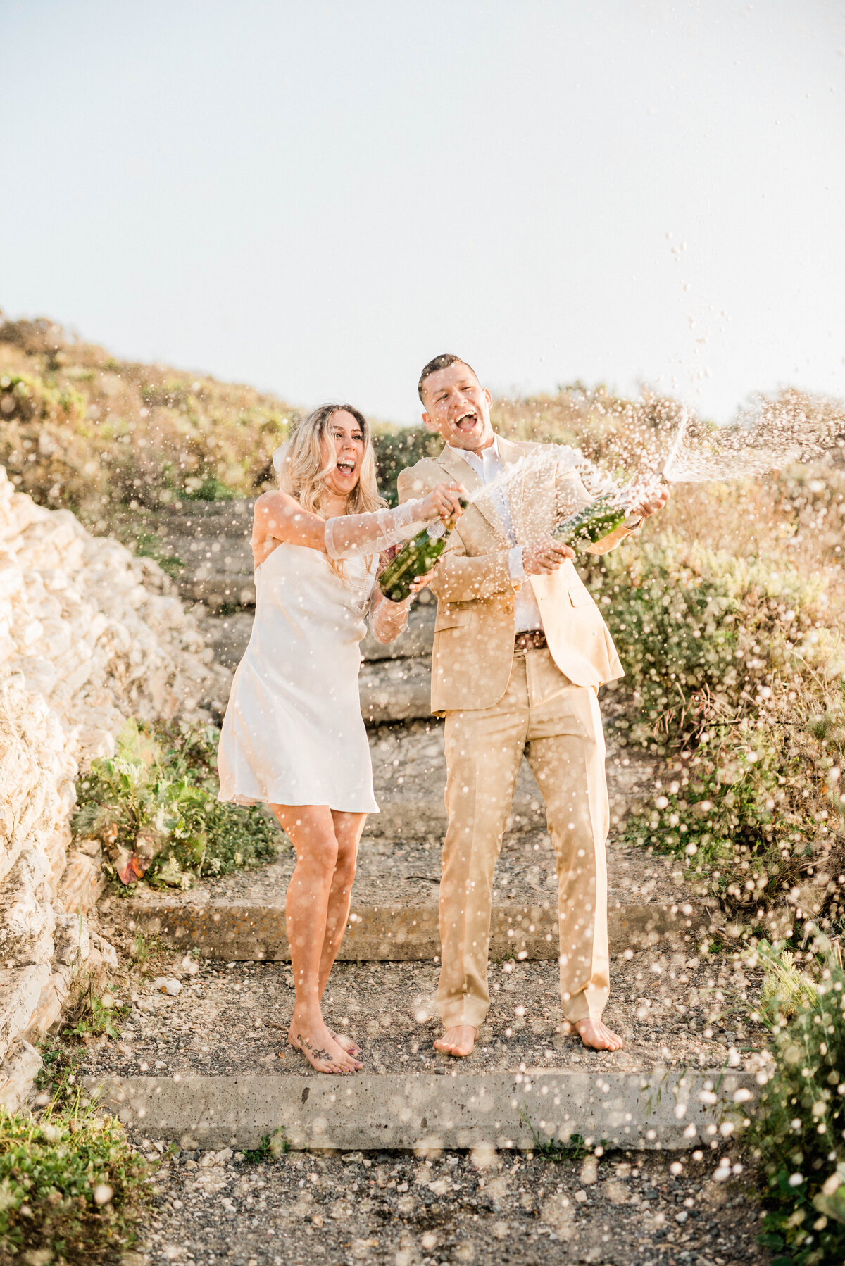 Engaged couple open a bottle of Champagne for a toast at their Montana de Oro engagement session. They happily spray Champagne and kiss.