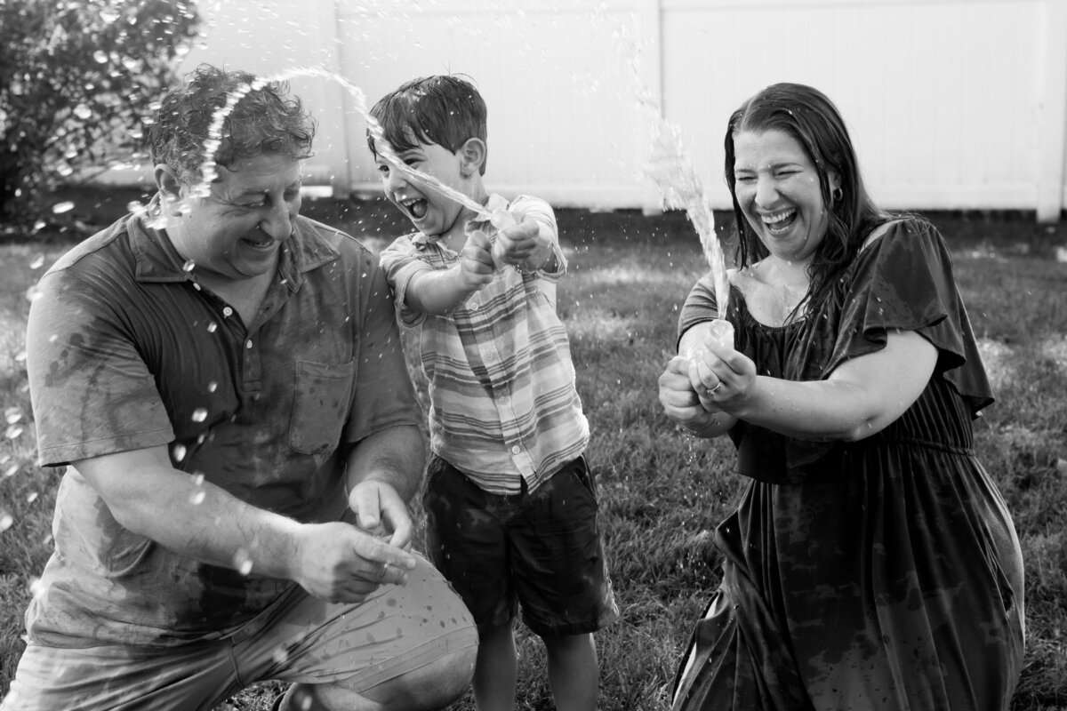 Black and white photo of. family of three have a water balloon fight in a family photo session  Hudson Valley family photographer Milestone Images
