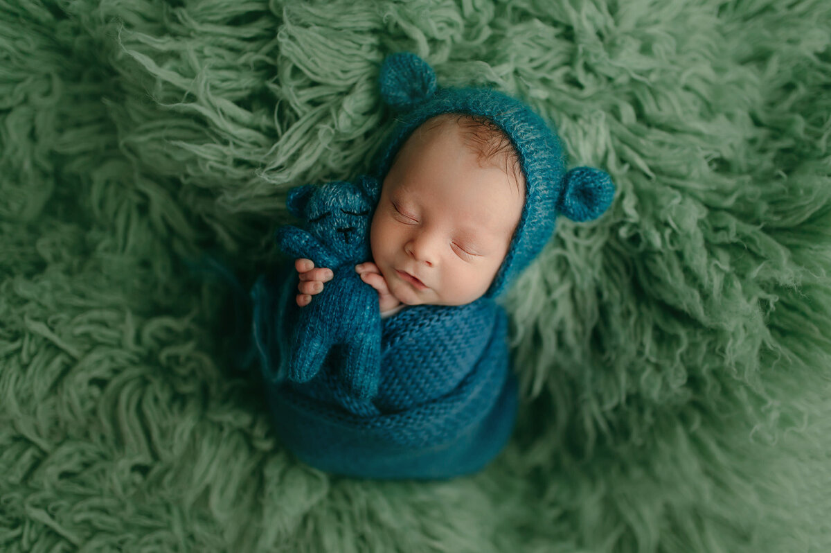 Portrait of a newborn baby wrapped in a teal, knit blanket and matching bear-eared hat. Baby is on a green, shag rug in our Waukesha, WI studio.