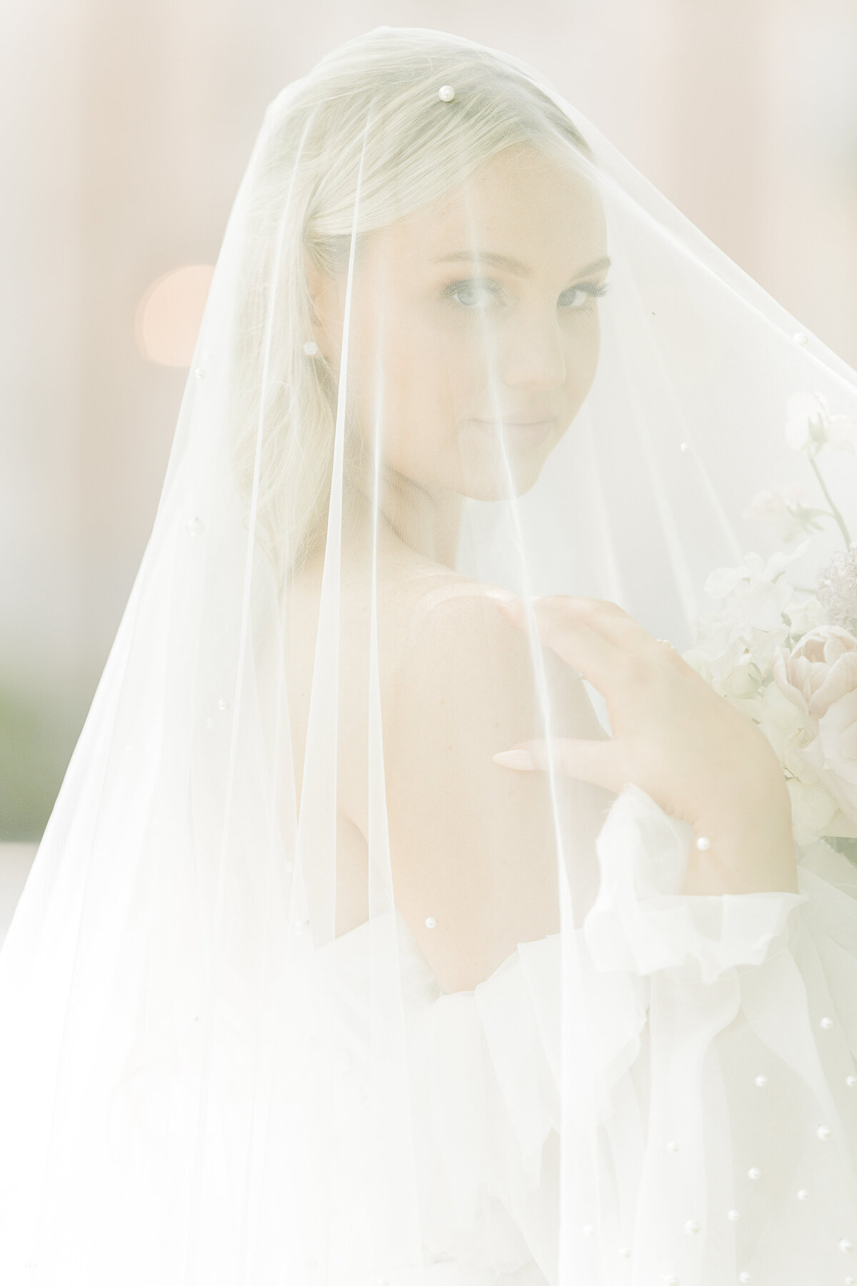 A North Shore bride poses for a formal wedding portrait in Boston. The bride has her veil pulled over her face, her forehand touching her shoulder and back hand holding her bouquet under the veil. The image is light and airy, almost with a light haze. Captured by best MA wedding photographer Lia Rose Weddings