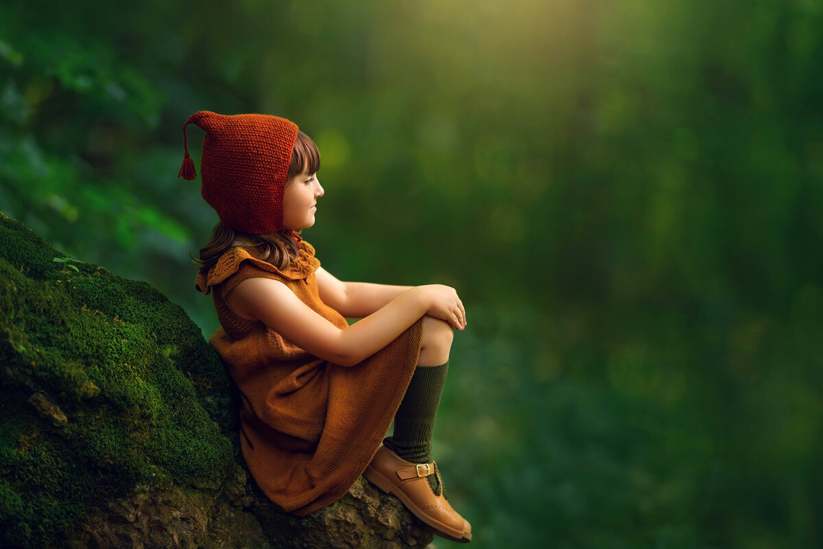 A young girl sits on a moss covered stone in hobbit attire for this whimsical portrait by Kara Reese Photography.