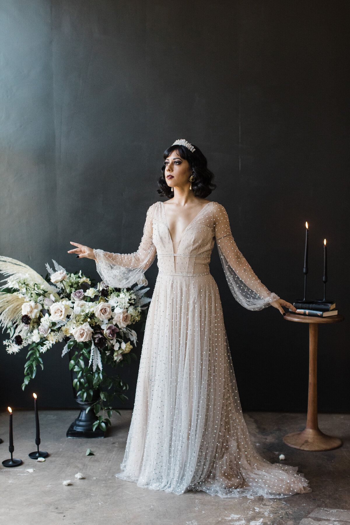 An artistic portrait of a bride on her wedding day in Dallas, Texas. She's wearing a long sleeve, flowing, intricate, white dress with a tiara and dark makeup. She's looking off to the side and reaching out her right arm in a focused yet relaxed pose. Her other arm rests on a small circular table covered with books and tall candles. On her other side are more candles and a large floral arrangement. The whole scene is backed by a black background.