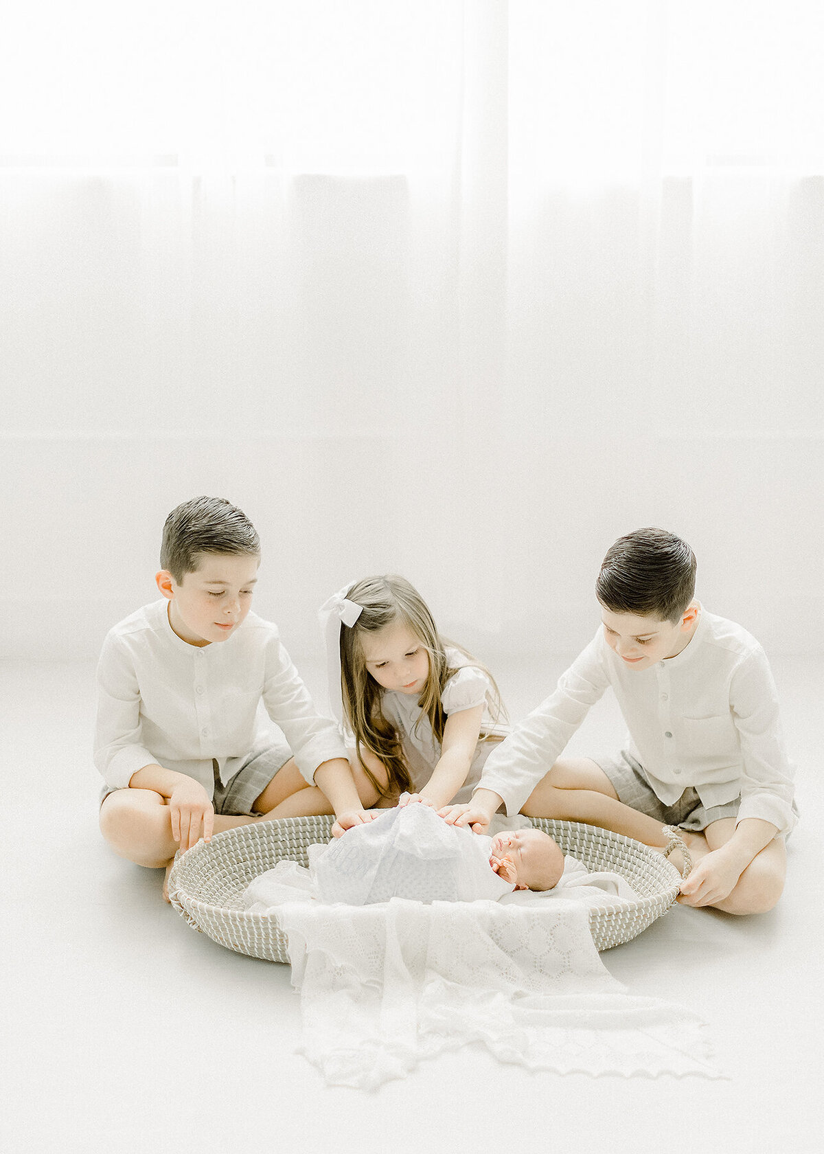 3 young siblings sitting on the floor of a Dallas newborn photography studio while their baby brother is swaddled and laying in a basket.