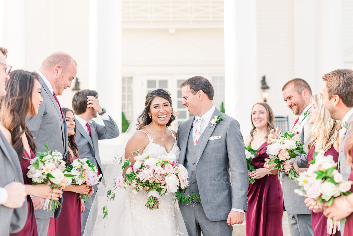 Smiling bride with her groom surrounded by bridesmaids and groomsmen in front of the Manor House.