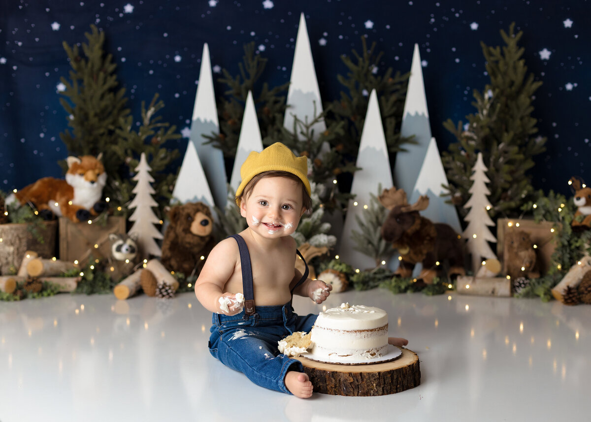 "Where the Wild Things Are" themed cake smash in top West Palm Beach and Jupiter photography studio. Baby boy is wearing jeans, suspenders, no shirt, and a gold crown against a backdrop of woodland creatures and a starry sky. Baby boy is smiling at the camera with icing on his hands and face.