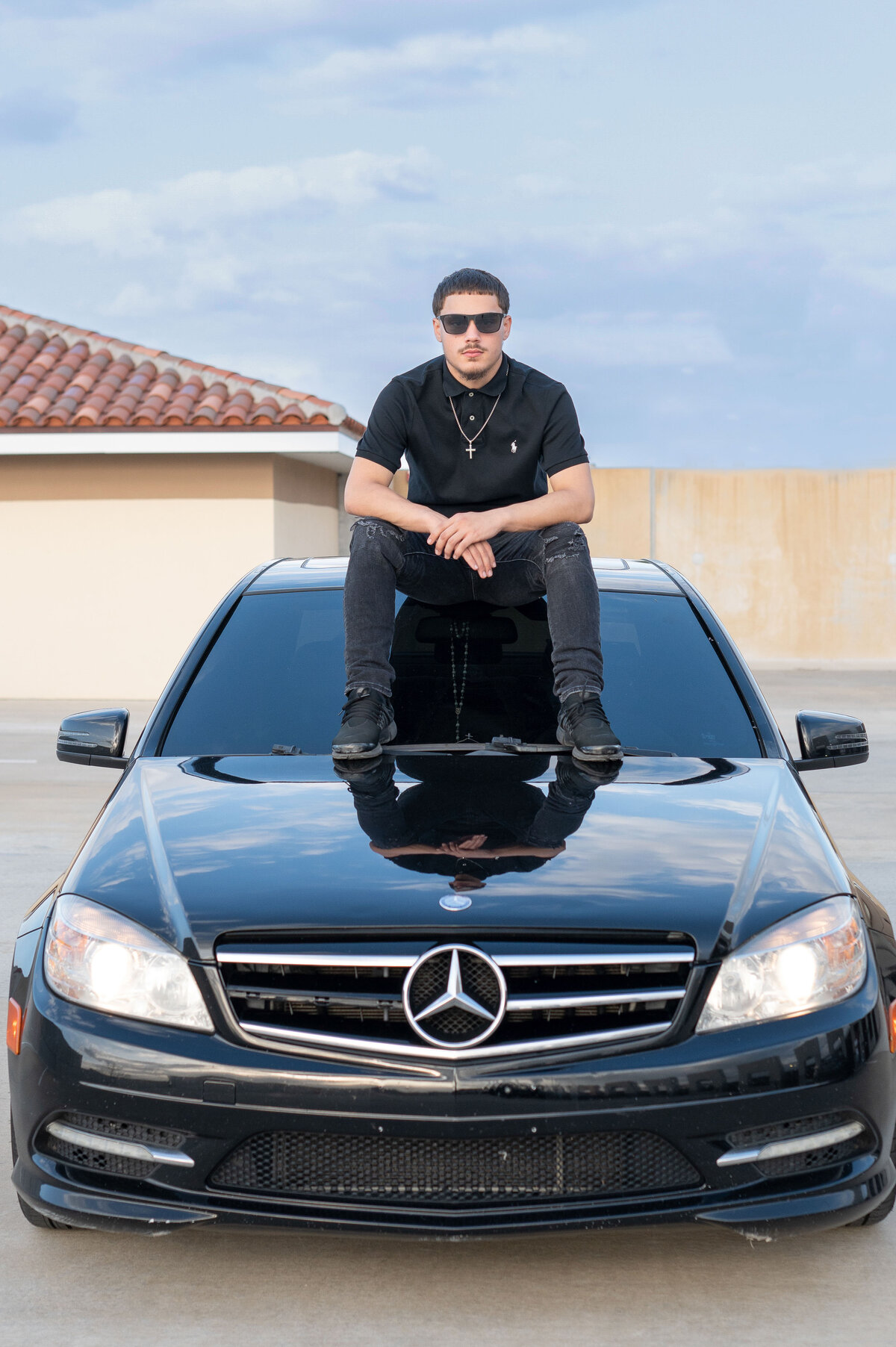 High school senior boy wearing sunglasses sits on top of black Mercedes car looking at the camera with a serious expression.