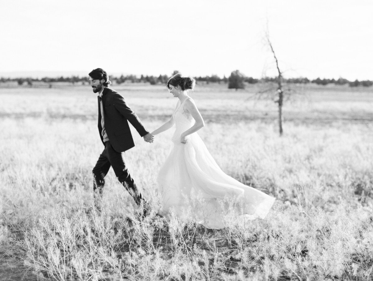 a black and white picture of a couple running through a field. he is hold her hand. she is in a white dress, hair pulled back and smiling.