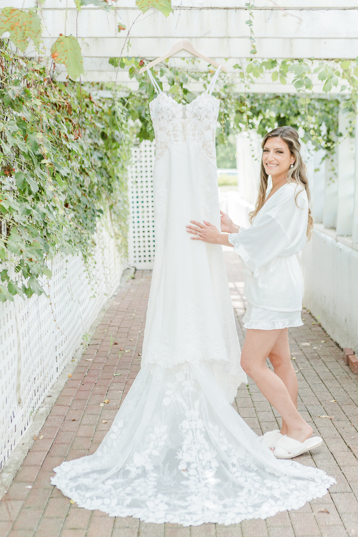 Bride stands beside her wedding gown hanging from the pergola at the Five Bridge Inn. Captured by best Massachusetts wedding photographer Lia Rose Weddings.
