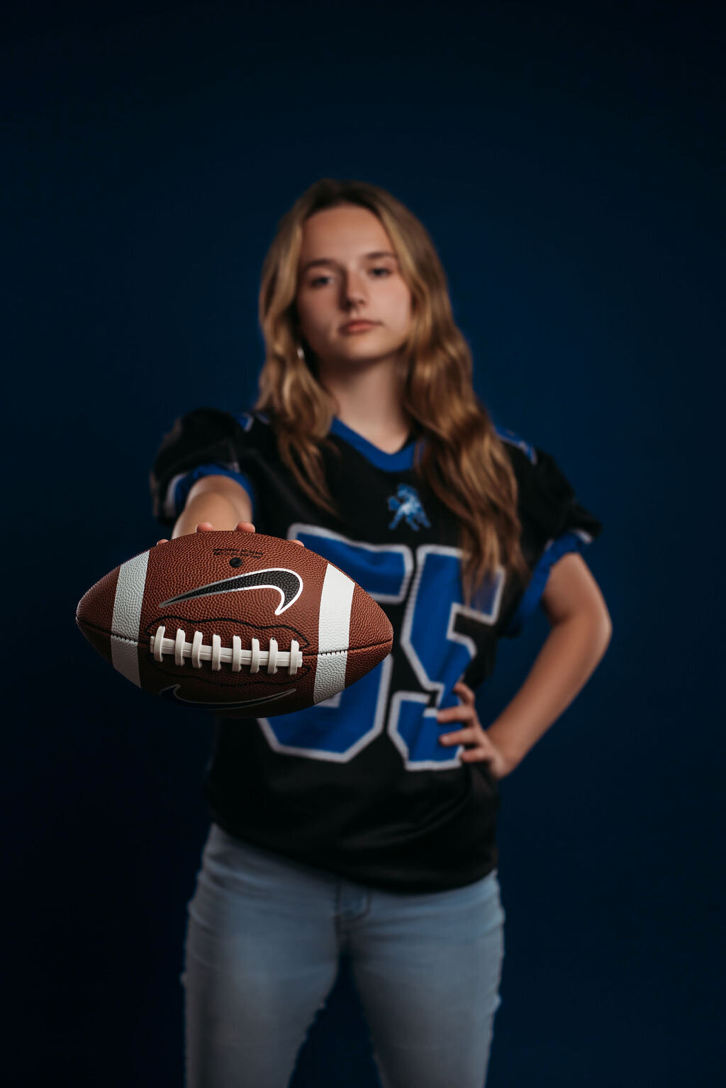 A teenage girl in a sports jersey holds out a football in front of a blue solid backdrop for her senior graduation photoshoot, captured by a fine art studio photographer in Bemidji, Minnesota