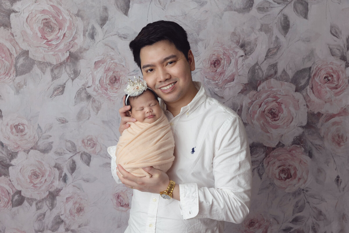 A happy new dad in a white shirt stands in a floral print studio holding his newborn daughter against his cheek