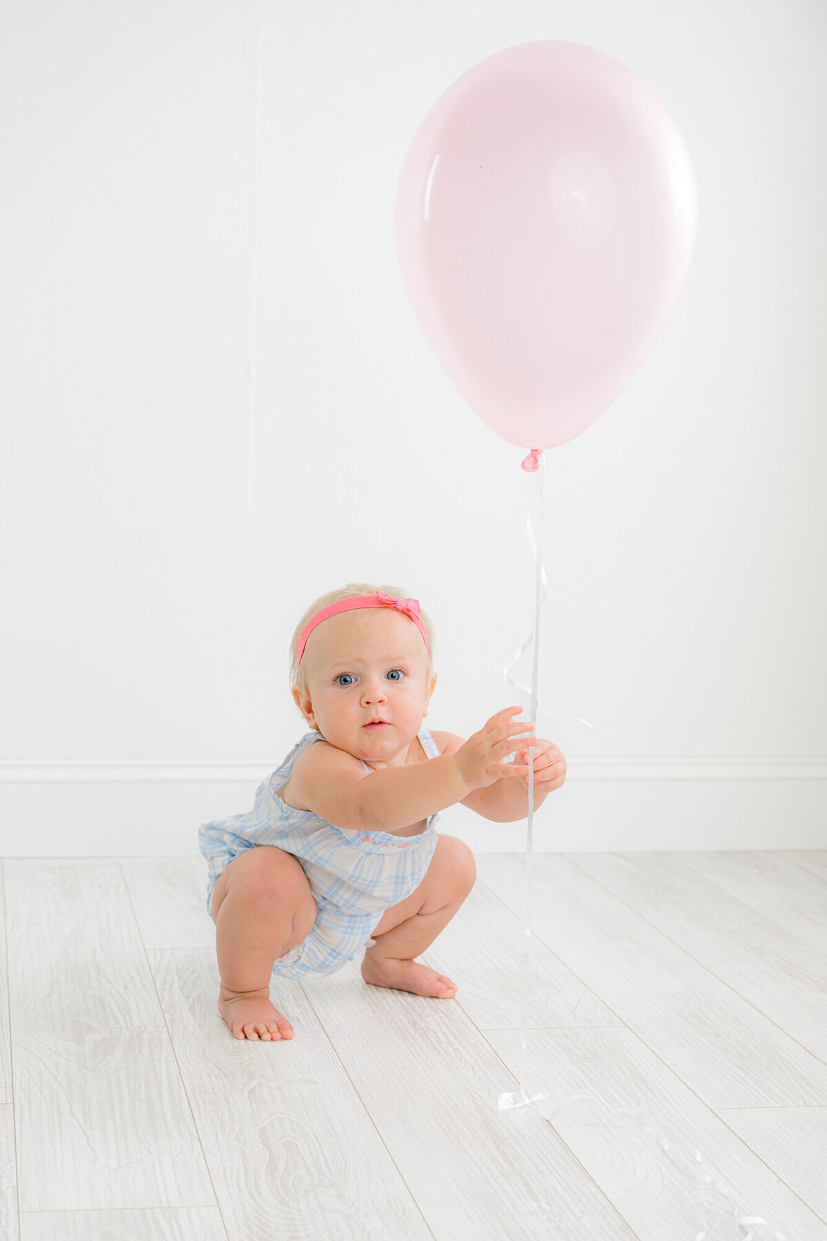 Little girls sits in a squat position holding a pink balloon as she laughs during her one year old Studio Milestone session