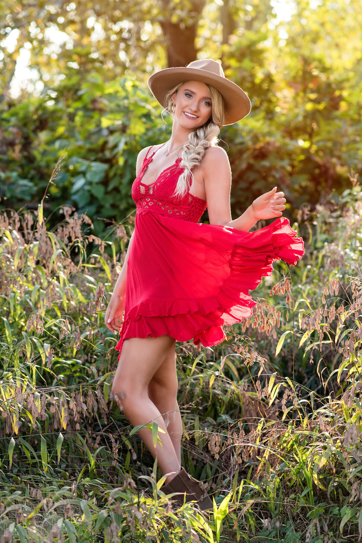 Clover Hill high school girl dances wearing red dress and tan hat in field of grass for her high school senior pictures.