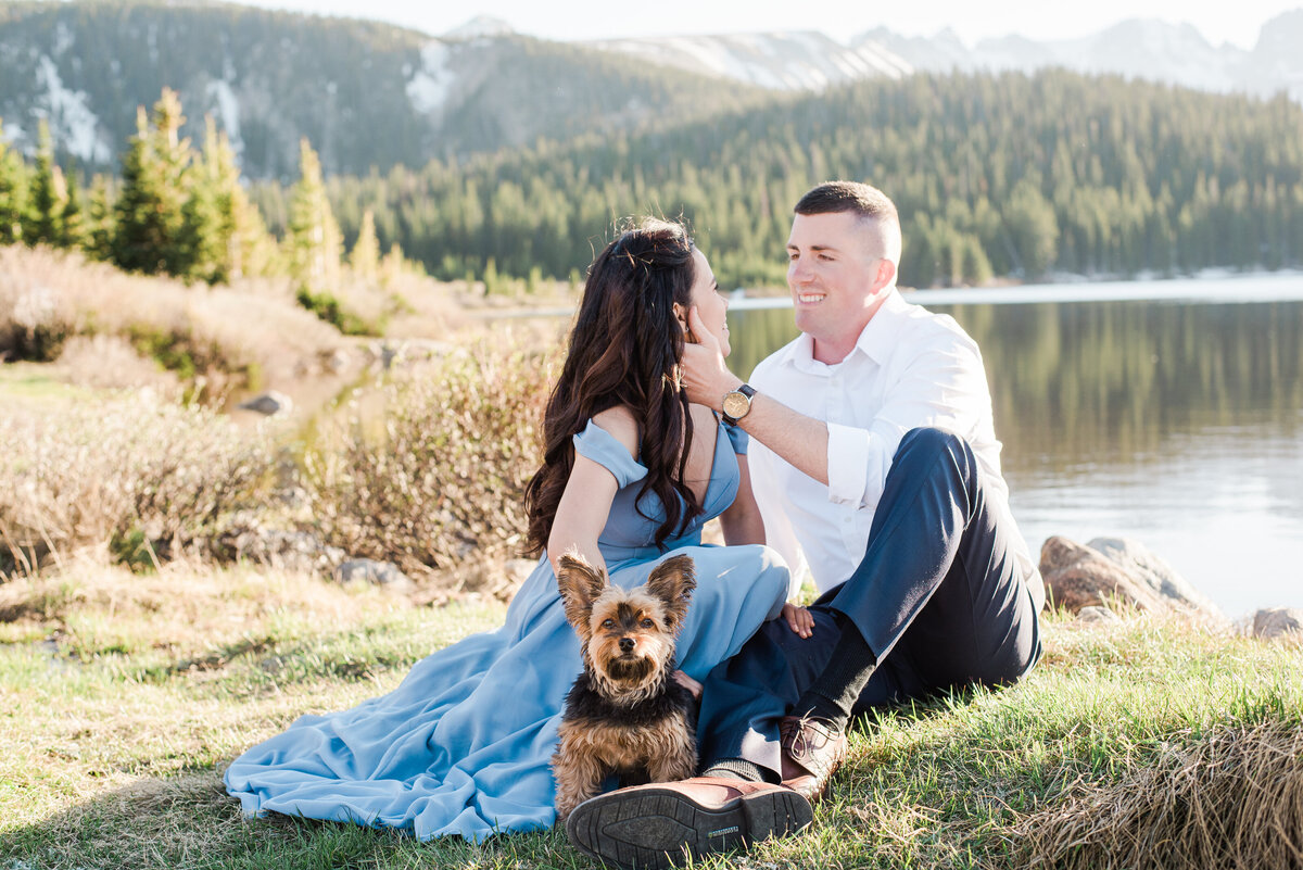 denver engagement photographer captures man and woman sitting together on a patch of grass next to a lake with the man caressing the woman's cheek as she looks back at him while holding her dog