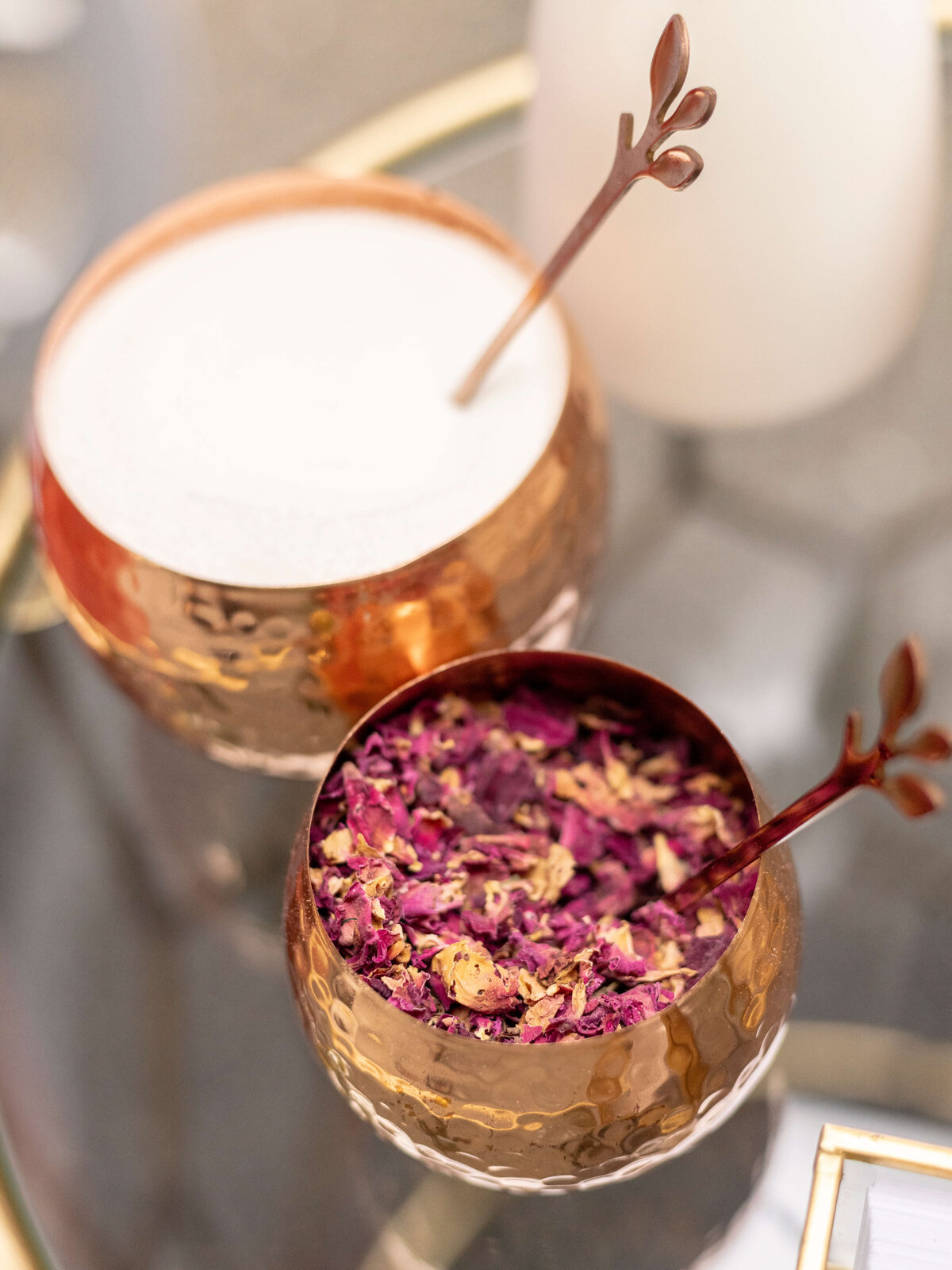 Two copper cups on a glass table, one filled with a creamy beverage and the other with dried rose petals. Each cup has a metal stirrer with a branch-like handle. The setting is elegant and refined.