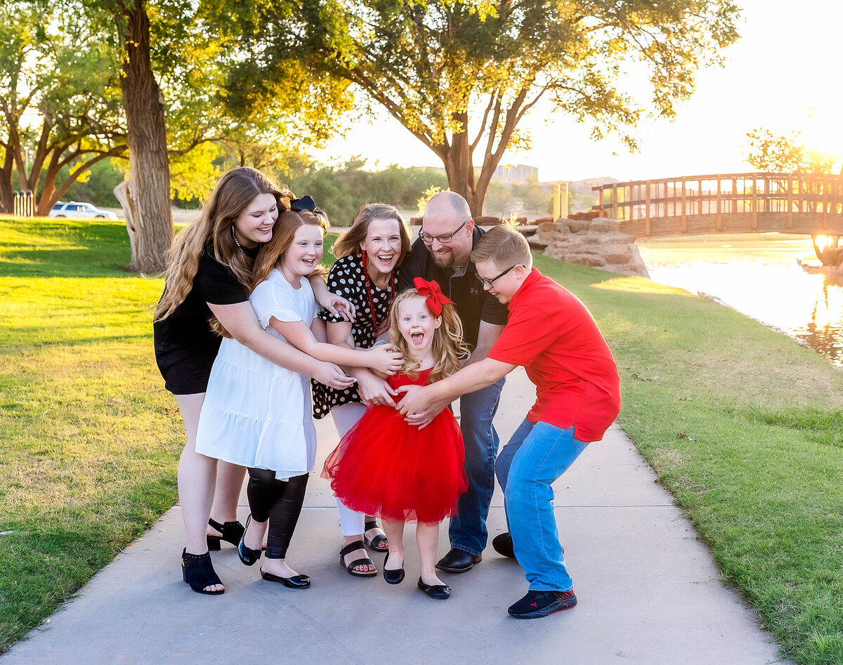 Family of 6 are dressed in red, white and black and are tickling the youngest child while she is laughing at the camera.