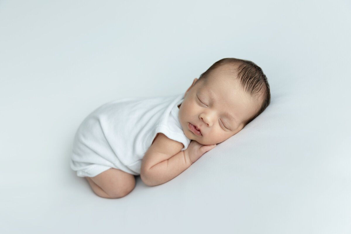 A newborn baby sleeps in froggy pose on a white bed in a white onesie in a studio