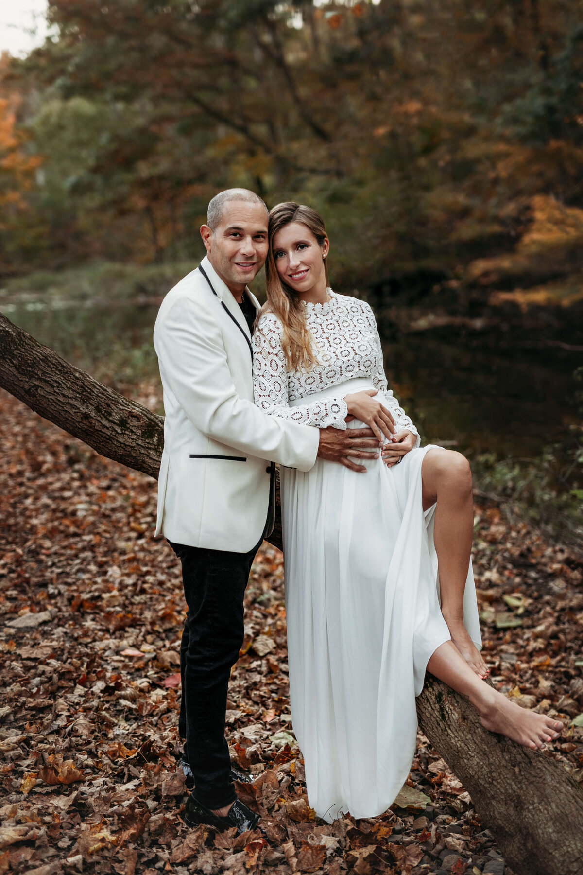 husband holding his pregnant wife who is wearing a white maternity gown and sitting on a branch in the fall