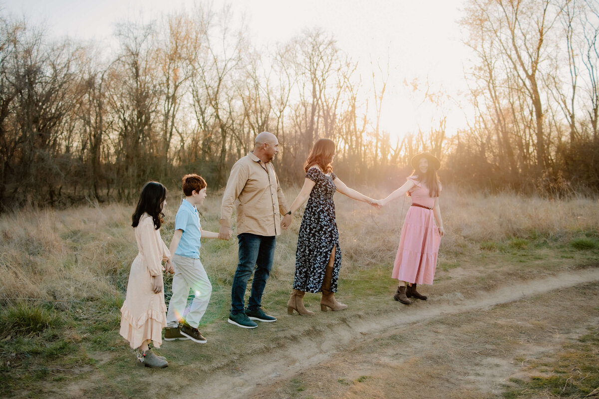 family of 5 walking up a slight hill of a dirt path  of a field holding hands and spread out  and the mom is looking back at the camera the daughter in the front of the line is looking back at mom