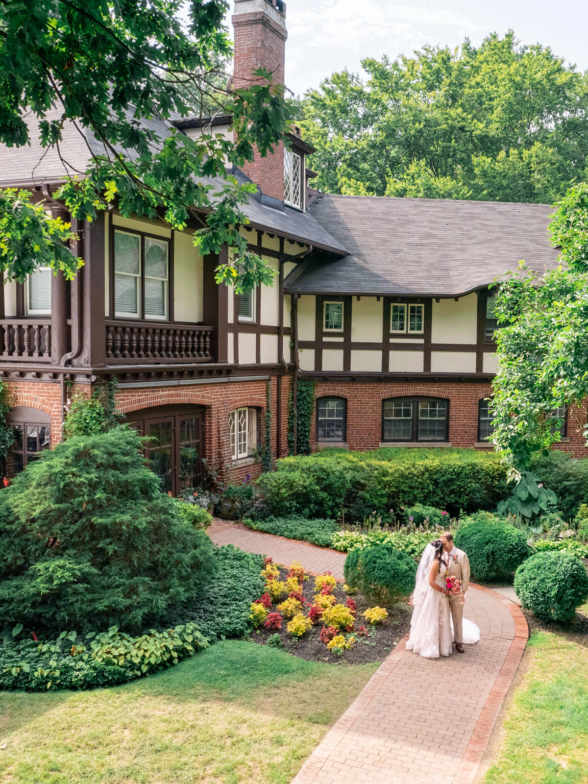 A couple in wedding attire stands on a pathway in front of a charming, two-story Tudor-style house surrounded by lush greenery and colorful flowers.