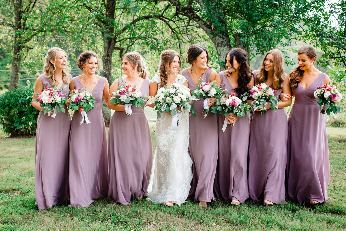 Bridesmaids in lilac dresses walking arm in arm with bride through field