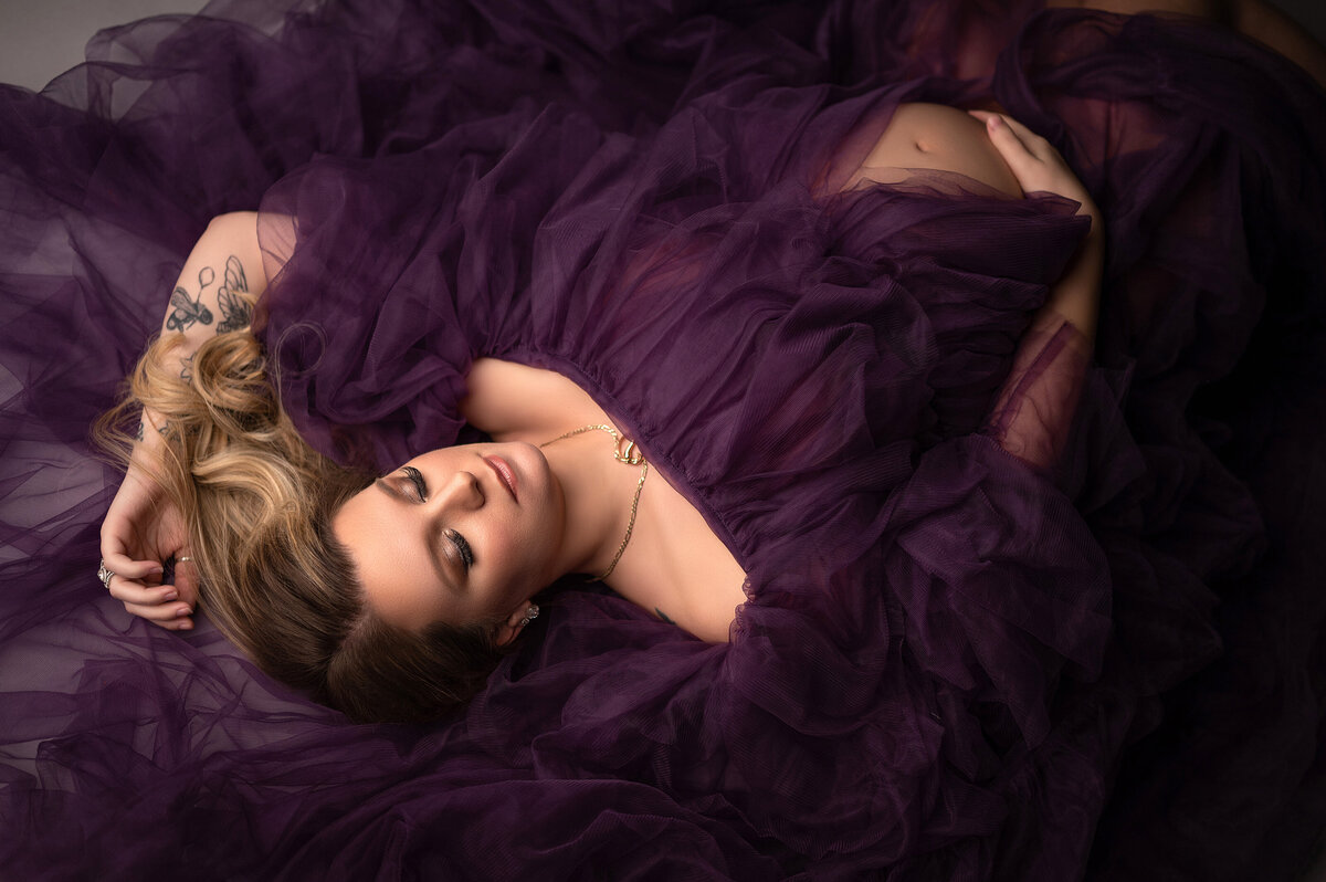 Pregnant woman lays in a  pile of sheer, purple fabric with her belly peeking through.  Her tattooed arm is raised above her head softly.