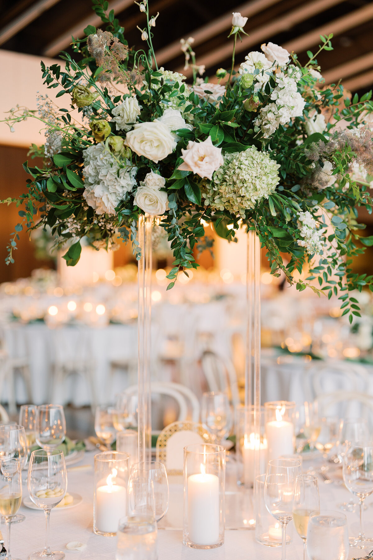Elevated floral centerpieces for garden-inspired wedding in downtown Nashville. Classic white and green wedding with floral colors in cream, white, taupe, and champagne. Timeless pillar and votive candles in white accent this lush tall floral design. Design by Rosemary & Finch in Nashville, TN.